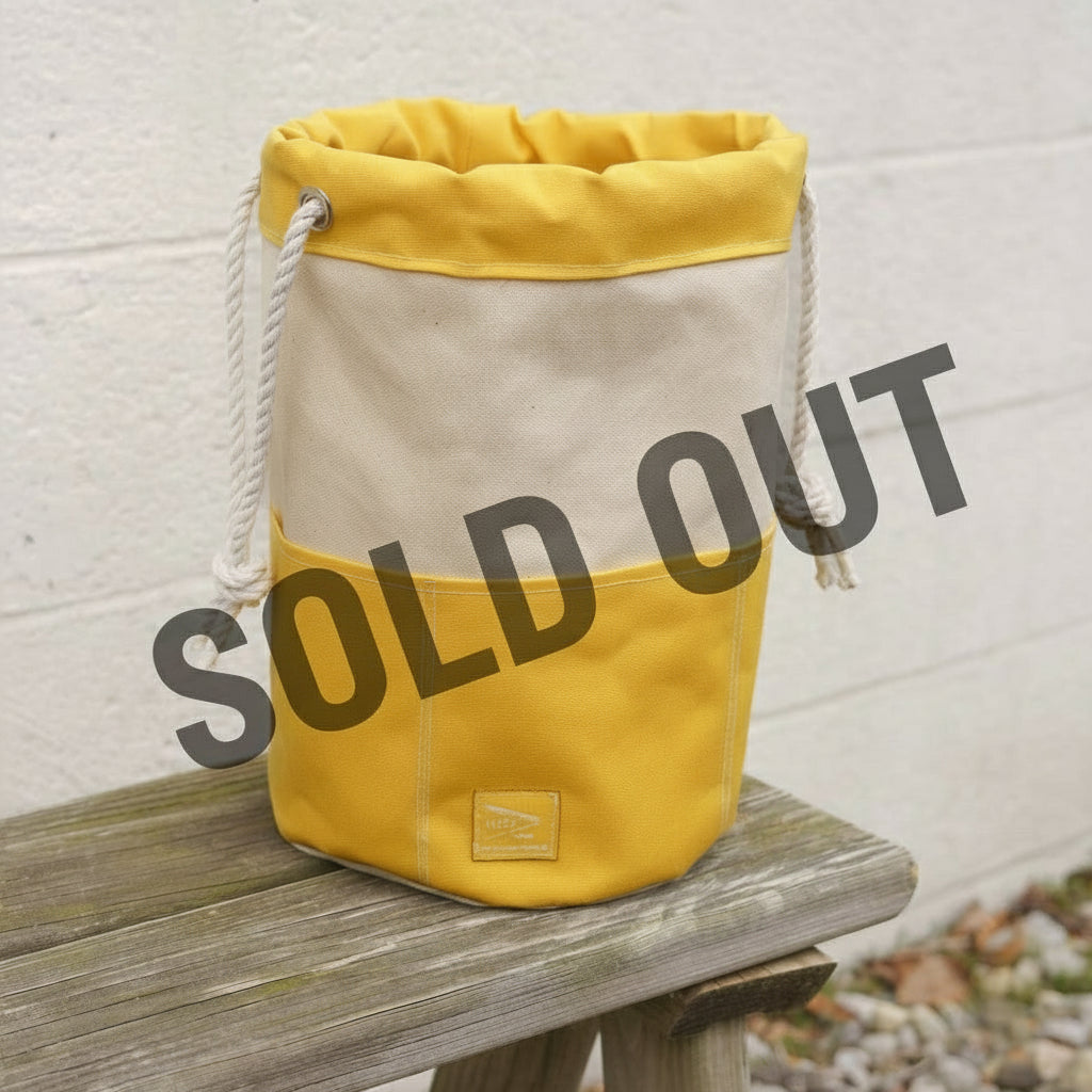 Yellow and beige bag with rope handle on a wooden surface against a light gray wall.