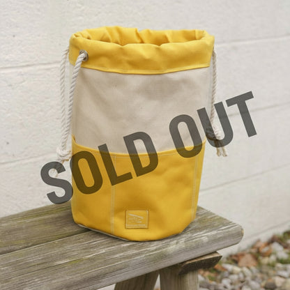 Yellow and beige bag with rope handle on a wooden surface against a light gray wall.